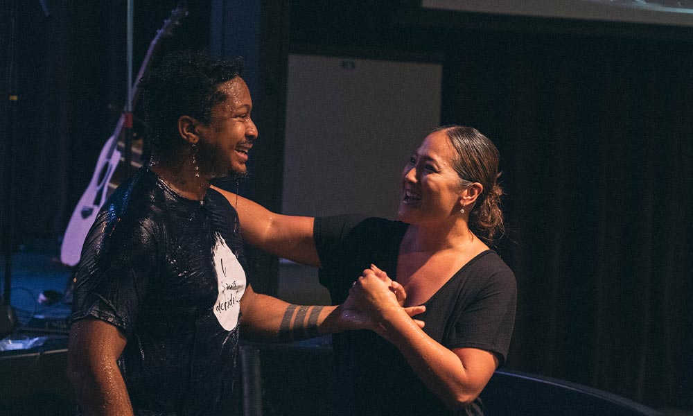A Black man stands in a baptismal, dripping wet and smiling. He has just been baptized by a Koreaan American woman pastor who is holding his hand and smiling at him.