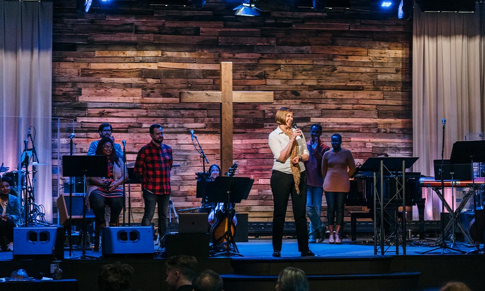A white woman holds a microphone and stands, smiling, on the Quest sanctuary stage. There is a wooden cross behind her and worship team members in the background