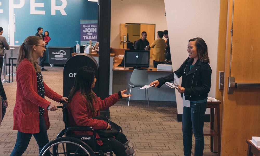 In the Quest main lobby, an Asian American woman smiles and hands a church bulletin to another woman in a wheelchair being pushed by her friend.
