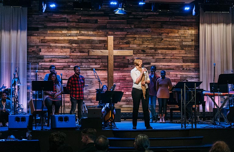 A white woman holds a microphone and stands, smiling, on the Quest sanctuary stage. There is a wooden cross behind her and worship team members in the background