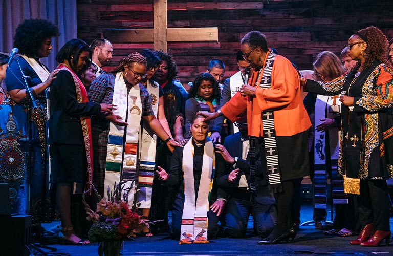 A group, mostly women of color pastors, gather around to lay hands on and pray over Pastor Gail Song Bantum, a Korean American woman. She is kneeling, her eyes are closed, and a hand is extended.