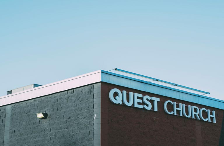 This is a photo of the outside corner of the Quest Church building - one side of the building is pained a dark red with the words Quest Church along the top. The other side is grey. The sky above is blue.